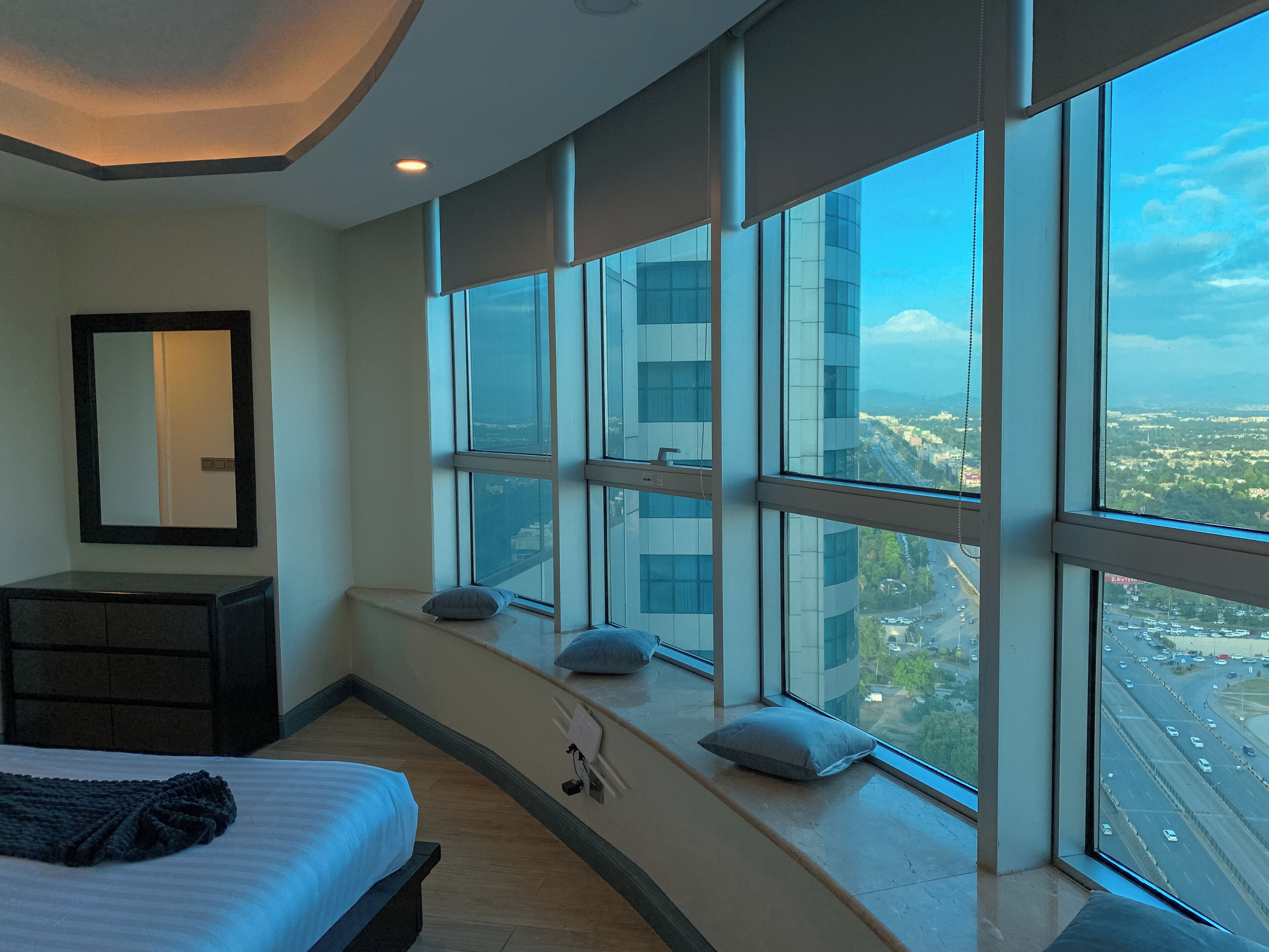 A modern hotel room with a large window, a bed, a dresser, and a mirror.
