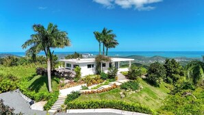 Une maison moderne à l’extérieur blanc, entourée d’une végétation luxuriante et de palmiers, et offrant une vue sur un paysage côtier.