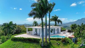 Une maison moderne avec une piscine, entourée d’une végétation luxuriante et de palmiers, offrant une vue sur les montagnes lointaines.