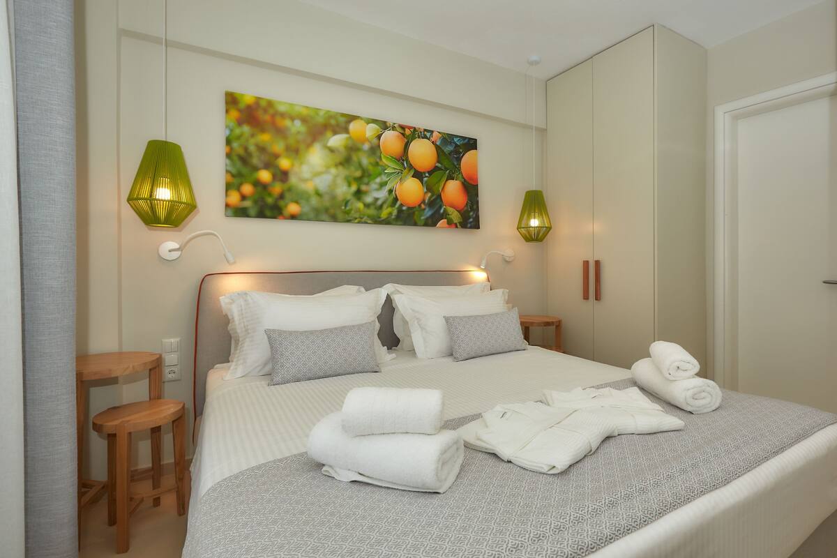 Un lit bien fait, avec du linge de lit blanc et à motifs, deux tables de chevet, un tableau représentant des oranges accroché au mur, et deux lampes murales.