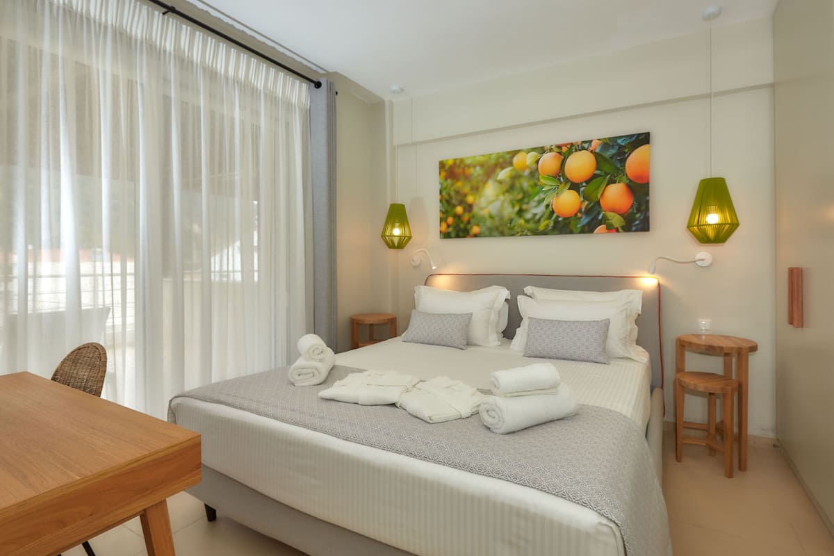 Un lit bien fait, avec du linge de lit blanc et une couvre-lit grise à motifs, deux tables de chevet avec des lampes, une chaise en bois et un tableau représentant des fruits accroché au mur.