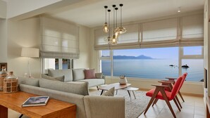 A modern living room with a sofa, a coffee table, and a dining area with chairs. There is a large window with a view of the sea and mountains.