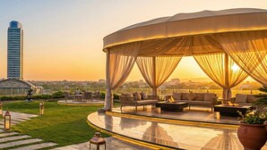 A rooftop terrace with a covered seating area, a view of three tall buildings, and a sculpture. - The Haven Residence (Islamabad)