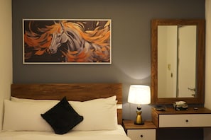 Une chambre d’hôtel avec une tête de lit en bois, un lit avec du linge de lit blanc et un oreiller noir, une table de chevet avec une lampe, un miroir et un tableau représentant un cheval accroché au mur.