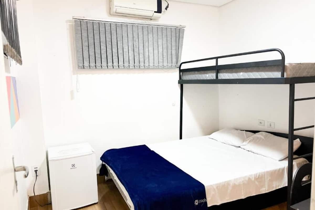 Une chambre avec un lit superposé, un petit réfrigérateur blanc, un radiateur et une fenêtre avec des stores.