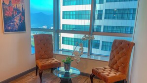 A room with two brown tufted chairs, a glass table, and a vase with white flowers. A large window offers a view of a modern building and mountains.