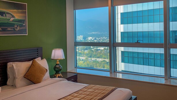 Habitación de hotel con una cama grande, una mesita de noche, una lámpara, un cuadro de un coche y vistas de la ciudad a través de ventanas amplias.