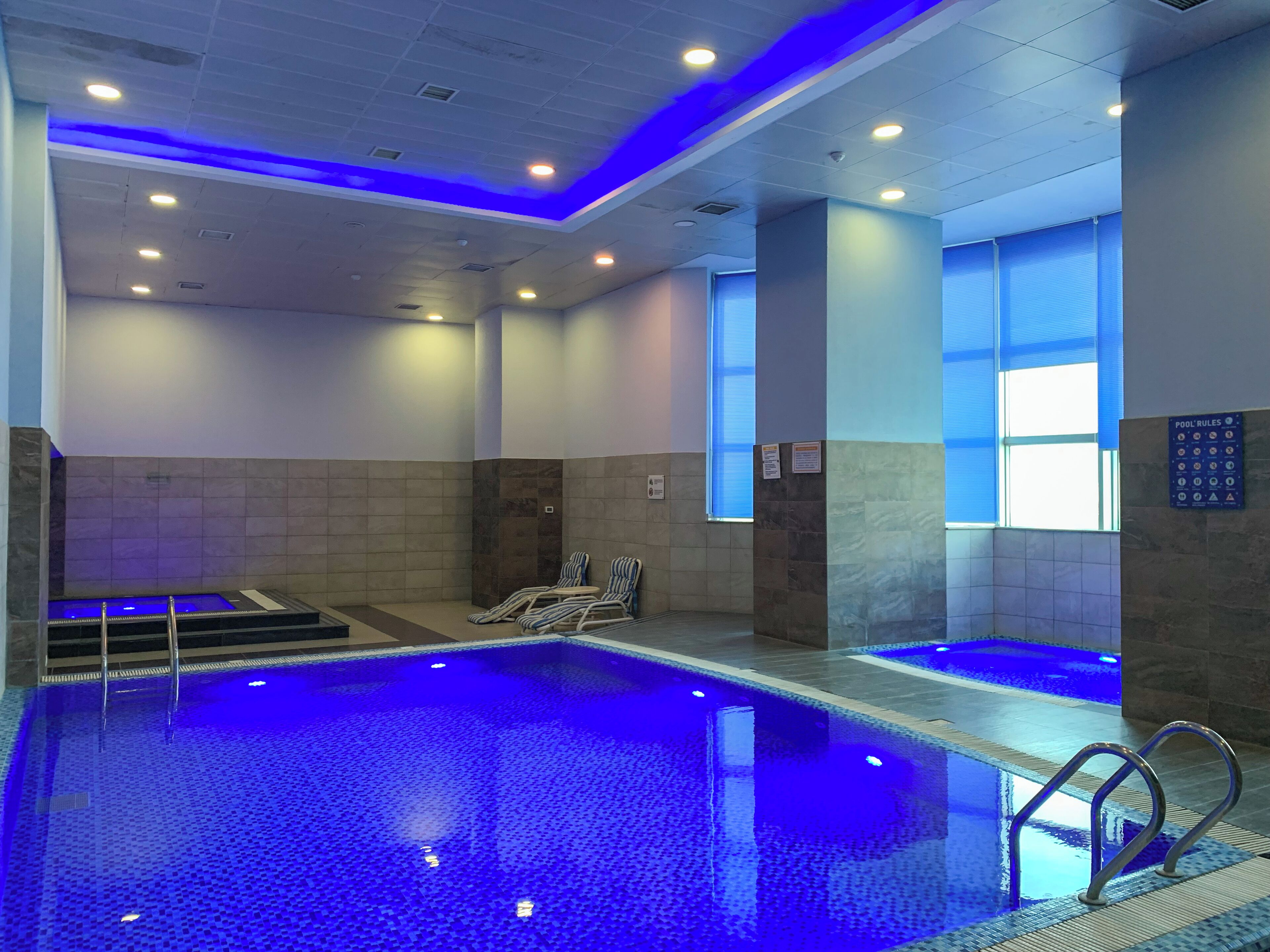Una piscina con iluminación azul, paredes con azulejos y una zona de borde con sillones de relax.