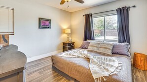 A bedroom with a bed, a ceiling fan, a window with curtains, a bedside table with a lamp, and a small wooden stool.