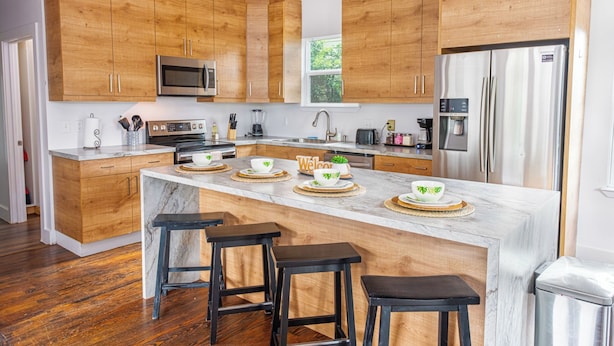 A modern kitchen with wooden cabinets, stainless steel appliances, a marble island with bar stools, and a view of the outdoors.