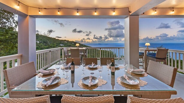 A dining table set for six with wicker chairs, a view of the sea, and string lights. - The Villa Michelle•Luxury•Ocean Views•Heated Salt Pool•Outdoor Living (St Thomas)