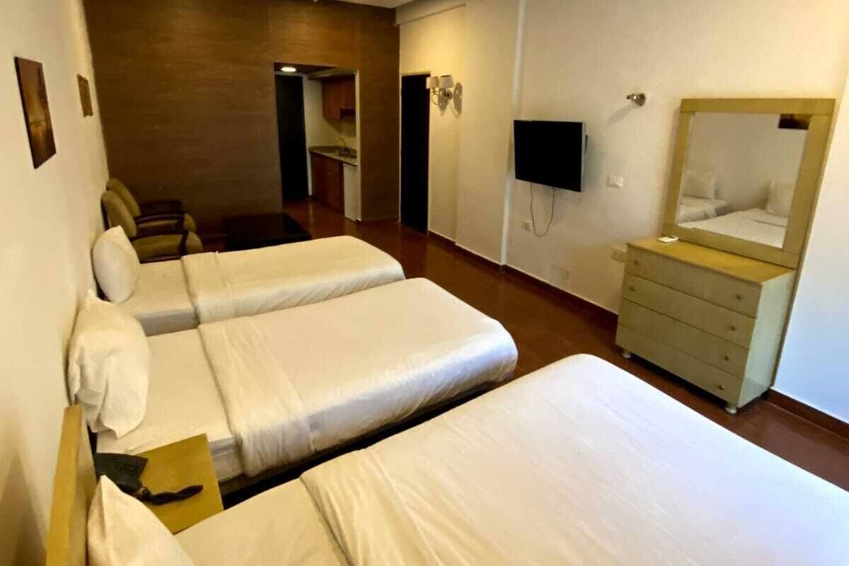 A hotel room with two single beds, a wooden dresser, a flat-screen TV mounted on the wall, and a small kitchenette area.