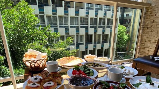 A balcony with a variety of breakfast items including croissants, pastries, and a bowl of soup, with a view of a building and greenery in the background. - Dreams Suites (Beirut)