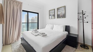 A modern hotel room with a large bed, a nightstand, a floor lamp, and a view of the city skyline through the window. - WelHome - Stunning High-Rise with Canal View Near MarinaMall (Dubai)