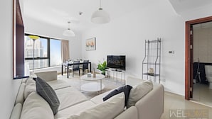A modern living room with a sofa, a coffee table, a TV, and a dining area. - WelHome - Stunning High-Rise with Canal View Near MarinaMall (Dubai)