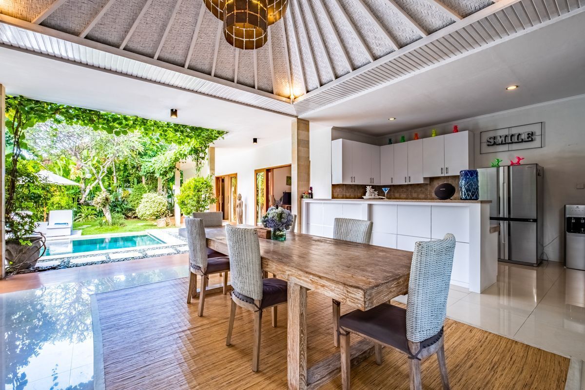 A spacious dining area with a wooden table and wicker chairs, a kitchen with white cabinets, and a view of a swimming pool and lush greenery.
