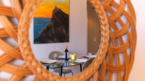 A dining area with a glass table, chairs, a bottle of wine, and a painting of a coastal landscape. - GuestReady - Idyllic vacation with stunning views (Caniço)