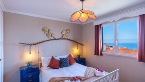 A bedroom with a bed, a nightstand, a blue dresser, a decorative branch headboard, a hanging light fixture, and a window with a view of the sea. - GuestReady - Idyllic vacation with stunning views (Caniço)