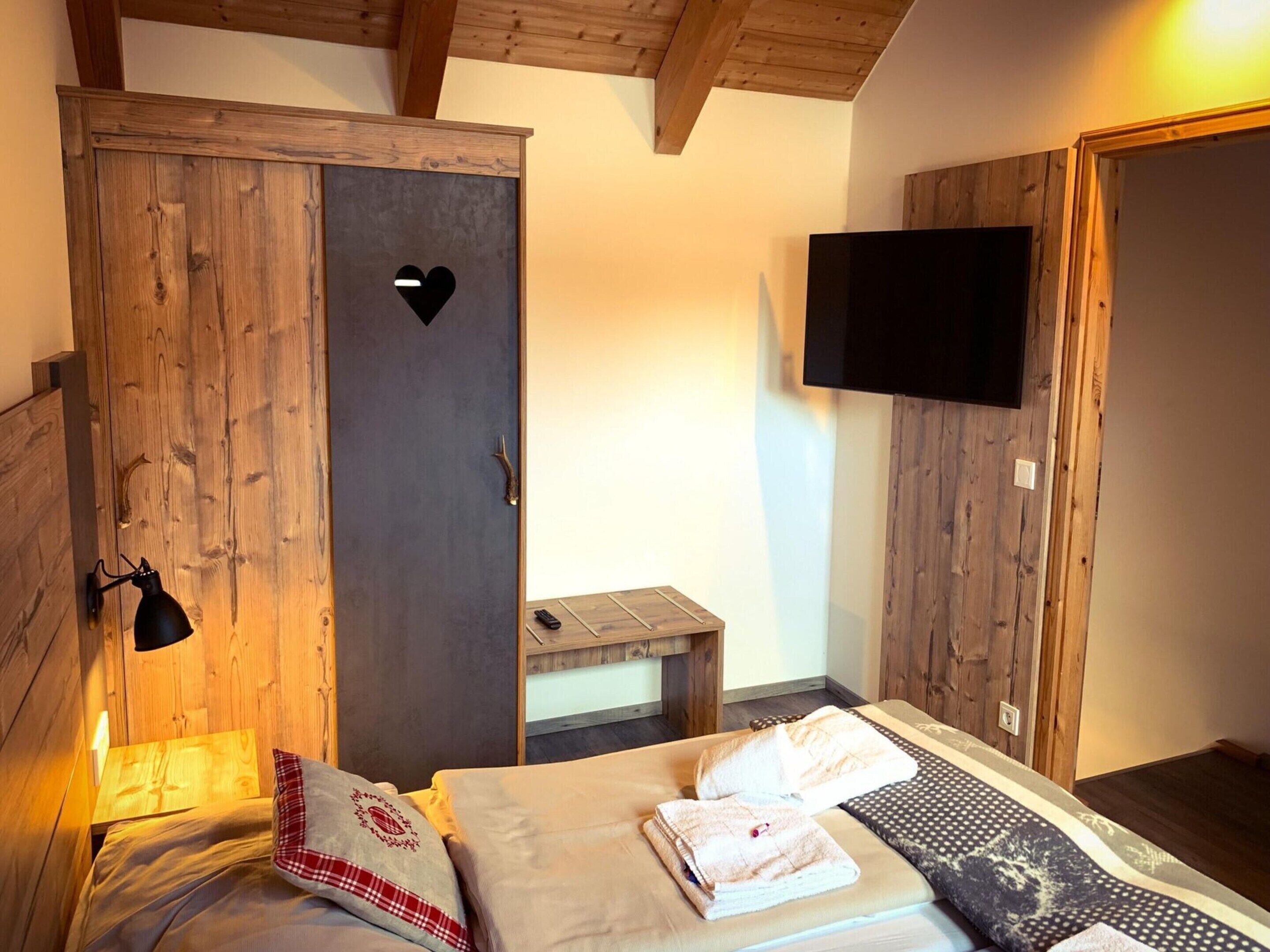 A bedroom with a wooden wardrobe, a bed with towels, a bedside table, and a mounted TV.