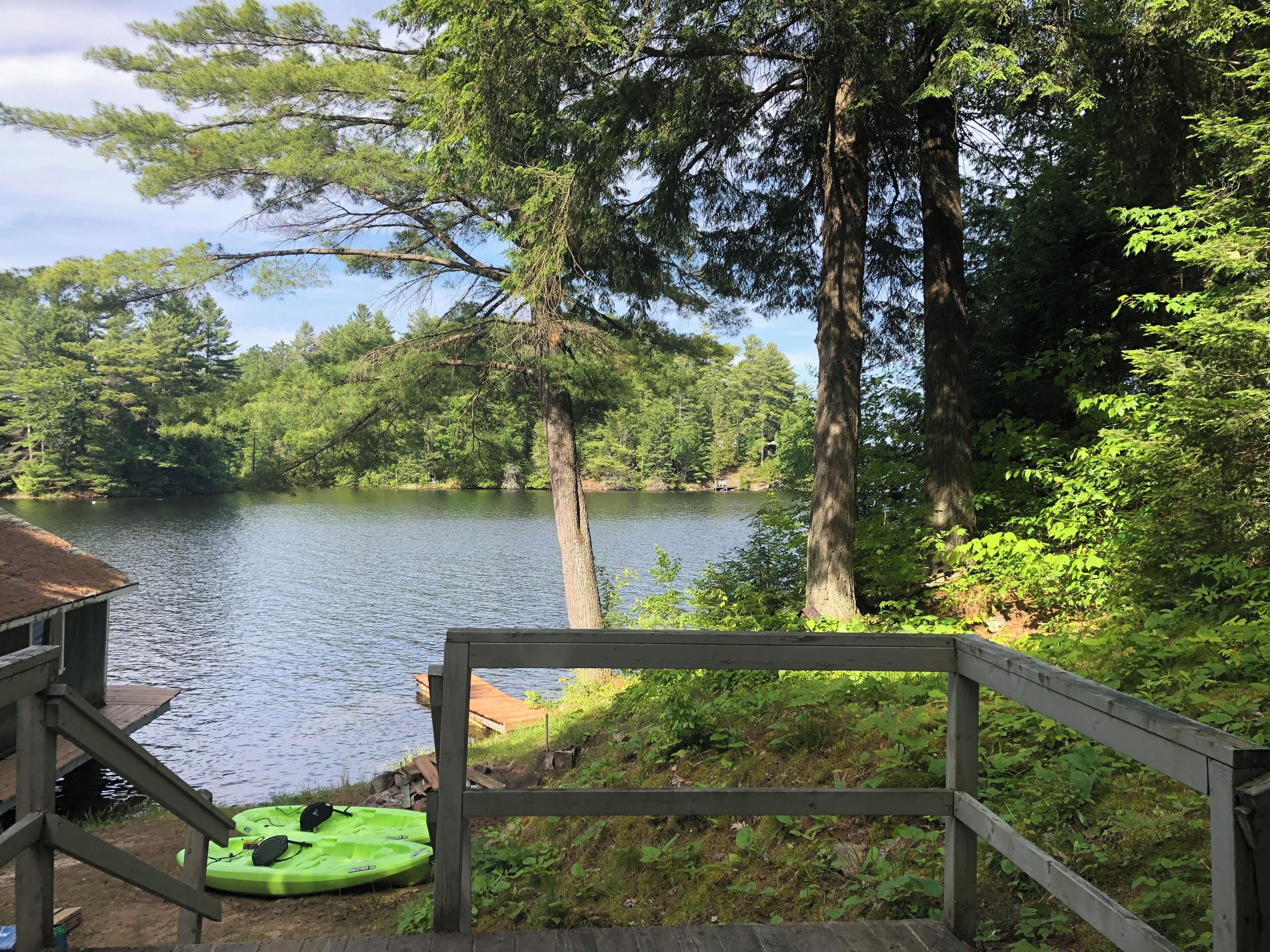 Eine Holzterrasse mit Blick auf einen See, mit einem Kajak und einem Stand-Up-Paddleboard, umgeben von dichtem Baumbestand.