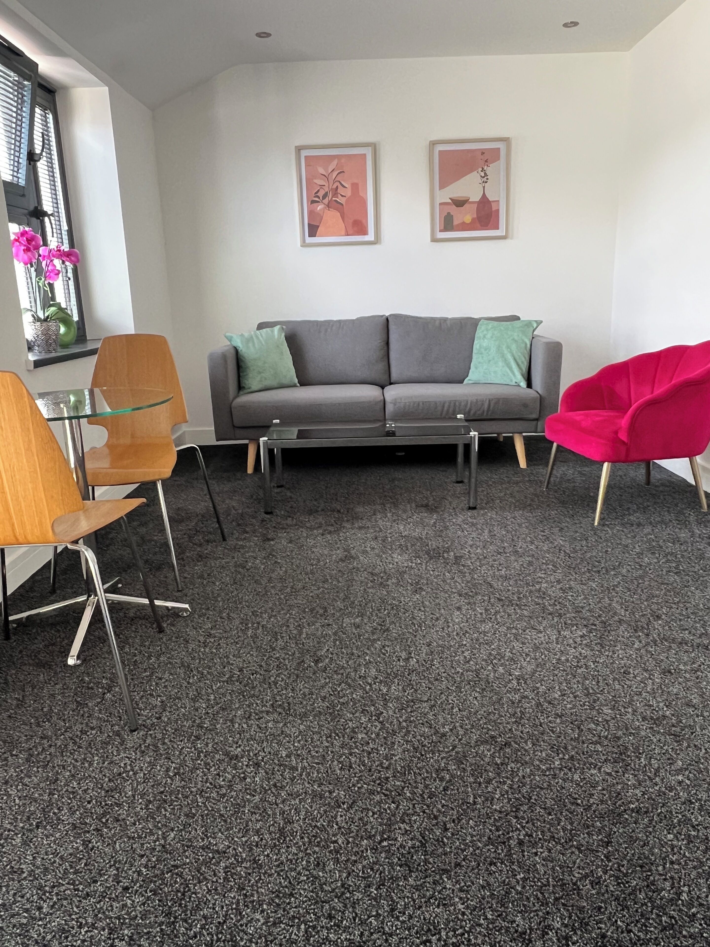 Ein modernes Wohnzimmer mit einem grauen Sofa, zwei grünen Kissen, einem Glastisch und einem roten Sessel. An der Wand hängen zwei gerahmte Kunstwerke und ein Fenster mit rosa Blüten.