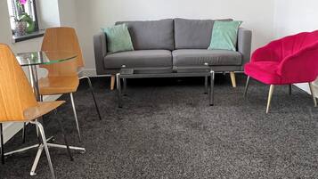 Ein modernes Wohnzimmer mit einem grauen Sofa, zwei grünen Kissen, einem Glastisch und einem roten Sessel. An der Wand hängen zwei gerahmte Kunstwerke und ein Fenster mit rosa Blüten.