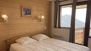 Un dormitorio con cabecera de madera, cama con ropa de cama blanca, dos lámparas fijadas a la pared y una ventana con vistas a las montañas.