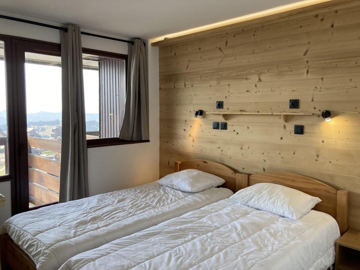 Cama doble con ropa de cama blanca en una habitación con paneles de madera y vistas a un paisaje urbano a través de amplias ventanas.