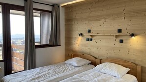 Cama doble con ropa de cama blanca en una habitación con paneles de madera y vistas a un paisaje urbano a través de amplias ventanas.