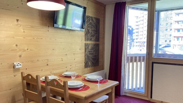 A wooden-paneled room with a dining table set for four, a television mounted on the wall, and a balcony with a view of buildings.