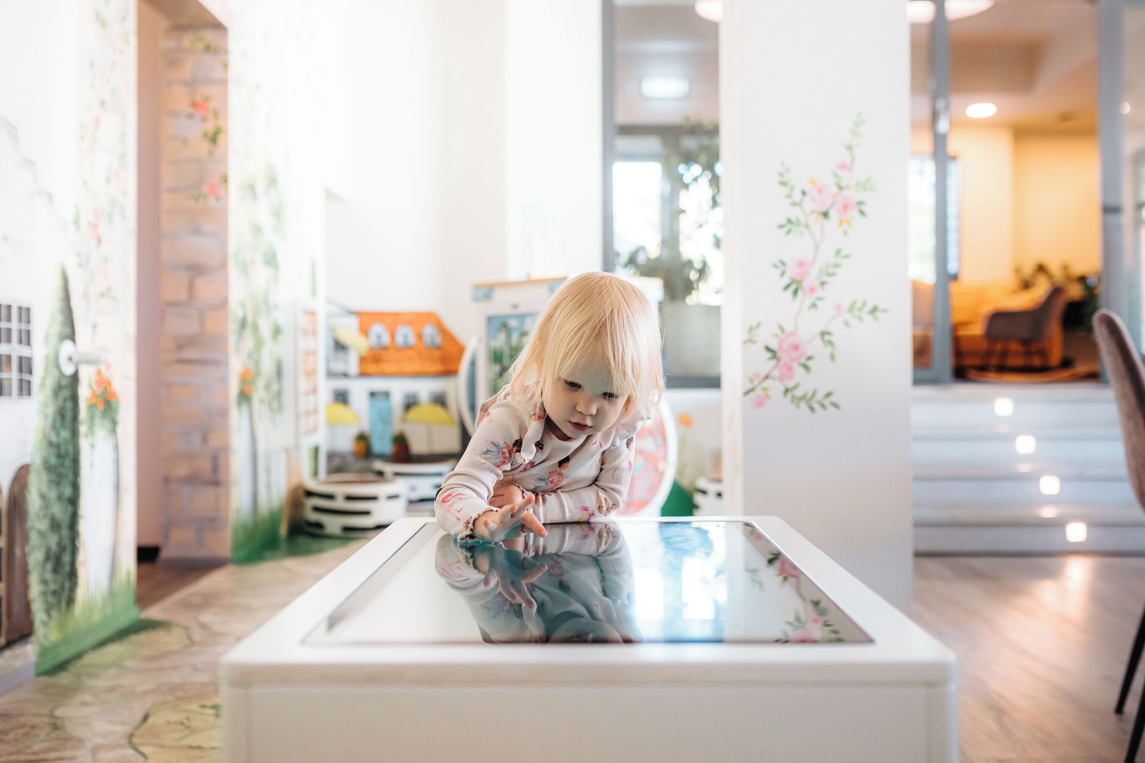 Un niño jugando sobre una mesa de vidrio en una habitación luminosa, con una chimenea y una escalera al fondo.