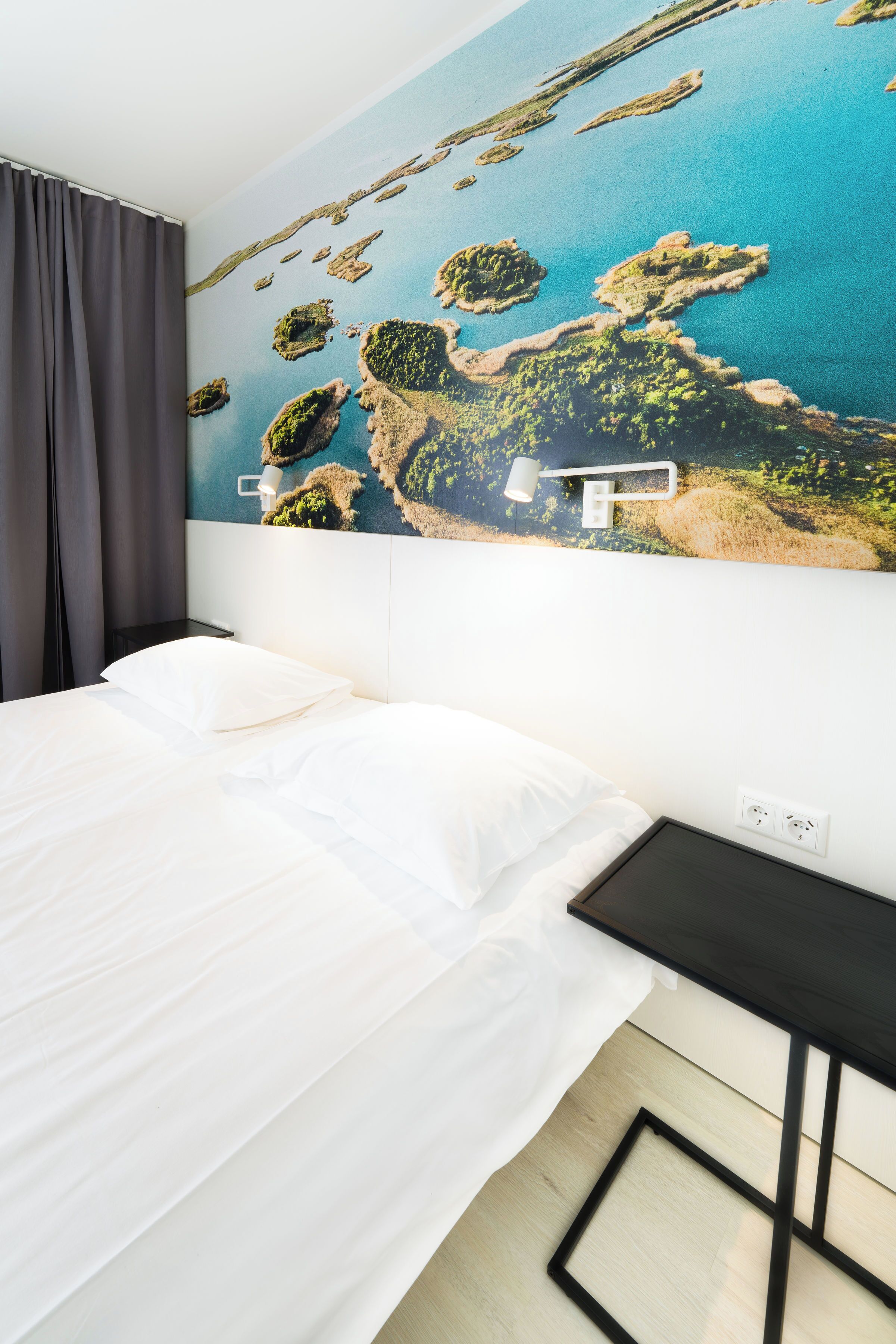 Un dormitorio con una cama, una mesita de noche, una lámpara de pared y un gran mural de un paisaje costero.