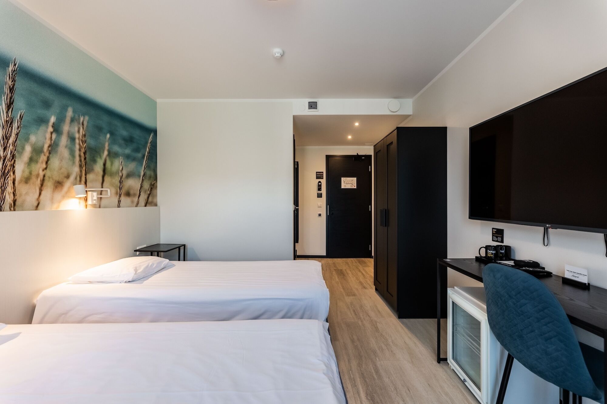 Quarto de hotel com duas camas, uma escrivaninha com televisão, uma mesinha e um quadro na parede.