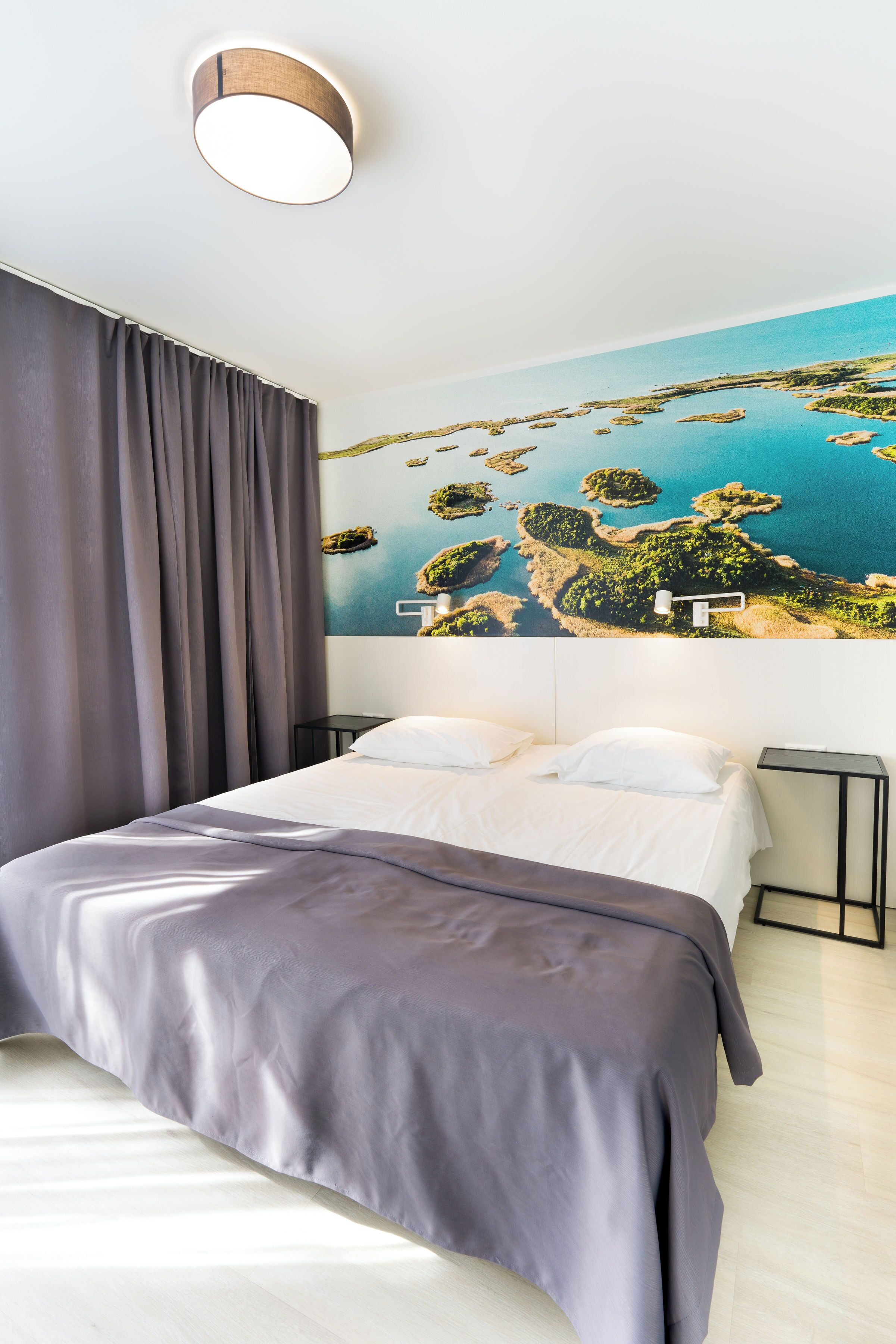 Una habitación de hotel con una cama grande, una mesita de noche, una lámpara y un mural en la pared con un paisaje costero.