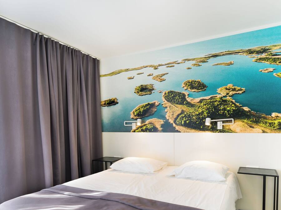Una habitación de hotel con una cama grande, una mesita de noche, una lámpara y un mural en la pared con un paisaje costero.