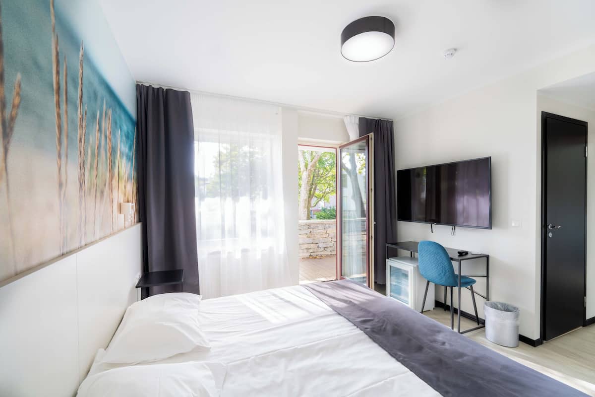 A modern hotel room with a large bed, a desk with a chair, a television, and a sliding glass door leading to an outdoor area.