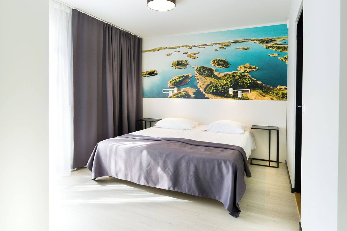 A modern hotel room with a large bed, a view of a coastal landscape, and a minimalist design.