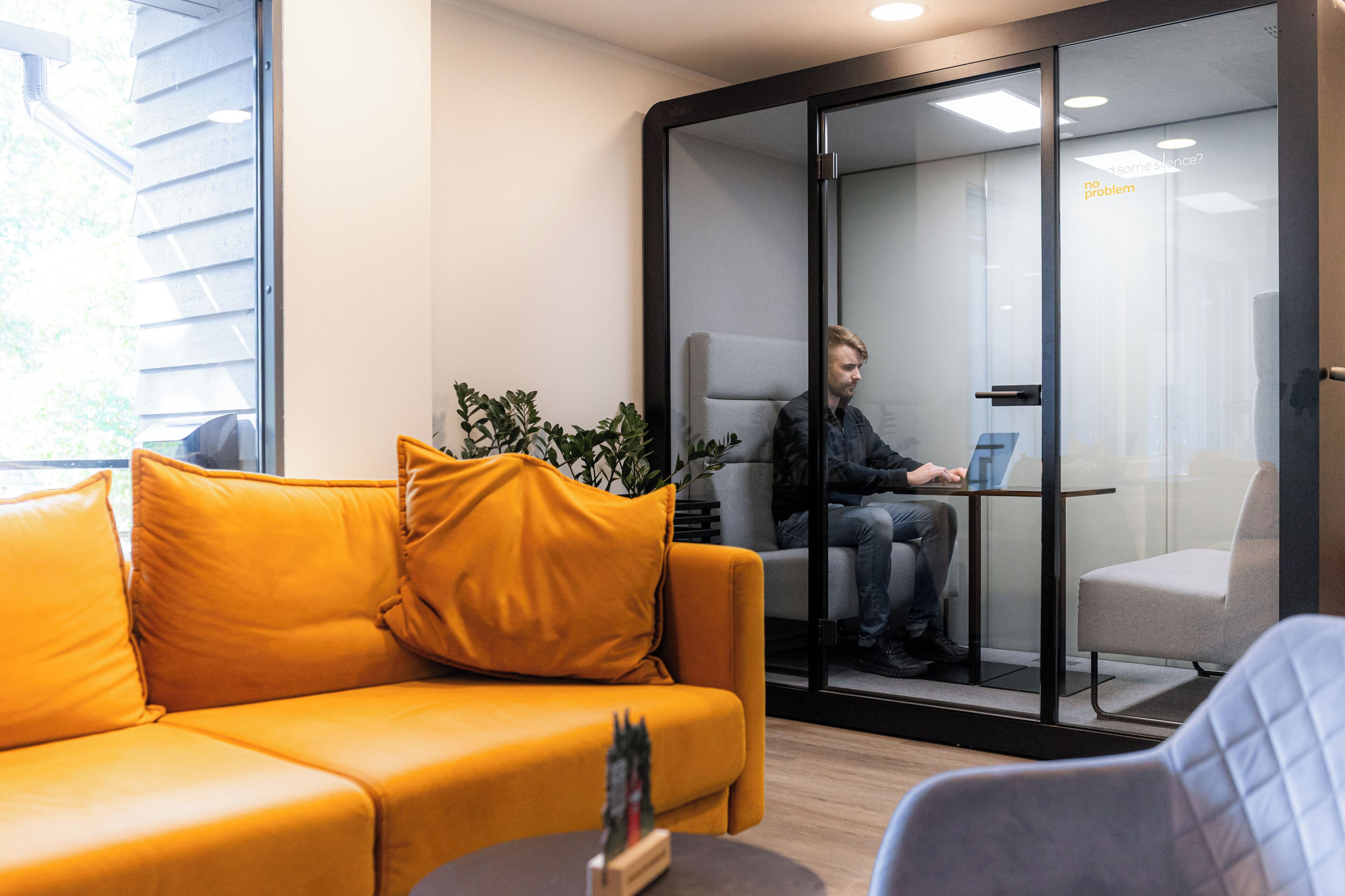 A modern office space with a glass partition, a yellow sofa, and a person working on a laptop.
