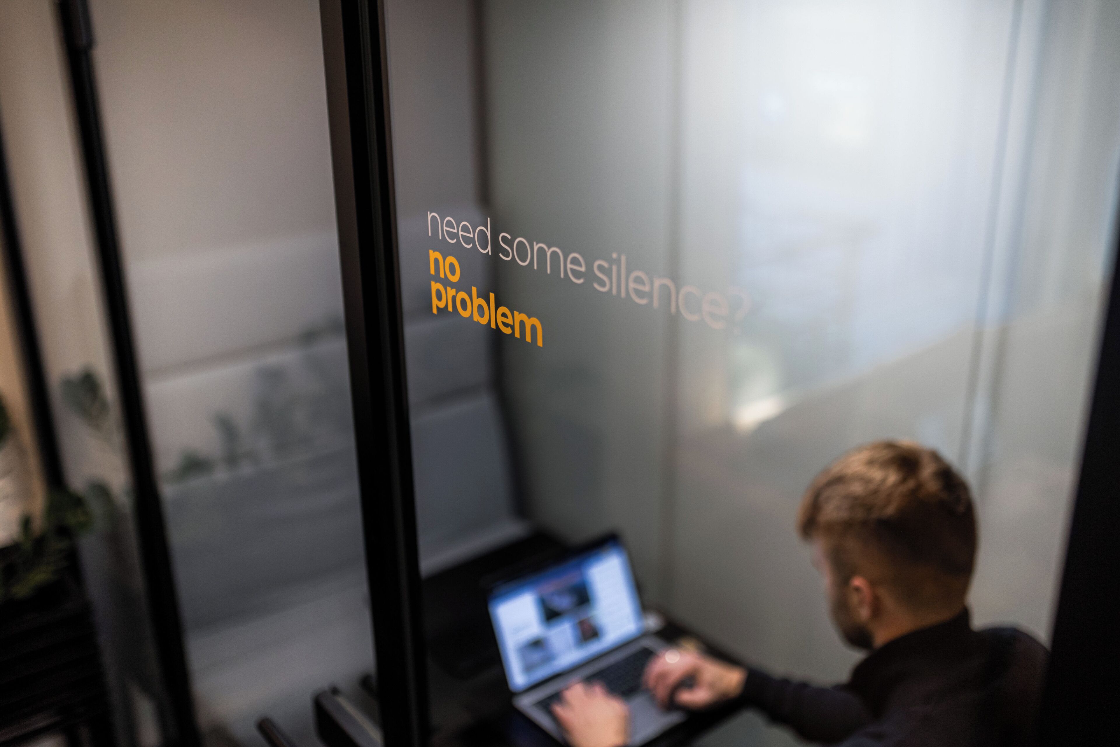 A person working on a laptop in a room with a glass wall displaying the text 'need some silence no problem'.