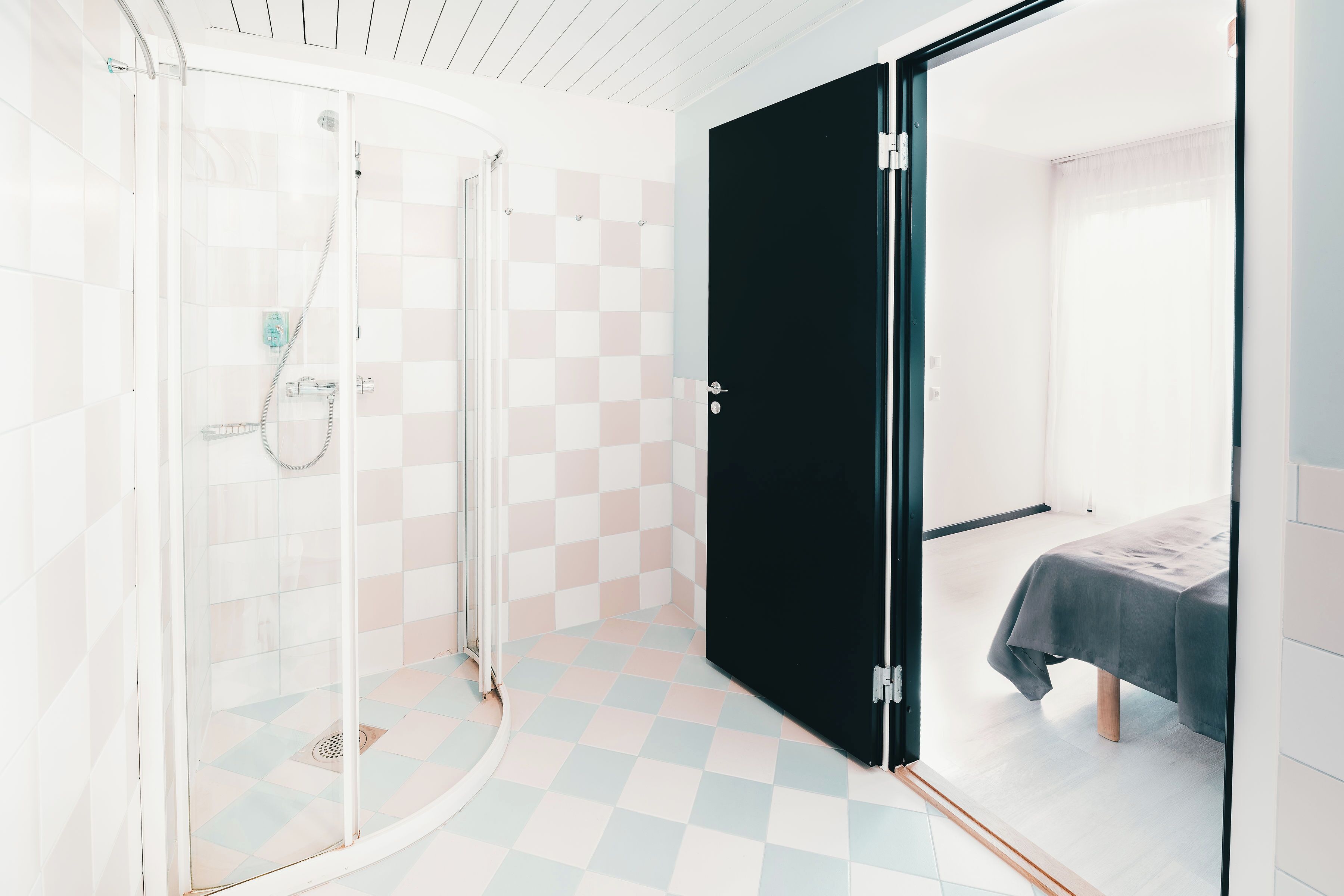Un baño moderno con cabina de ducha de vidrio, piso de cuadros y, en la habitación contigua, una cama con cubrecama oscura.