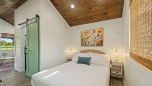 A bedroom with a bed, bedside tables, a green door, a painting, and a window with blinds.