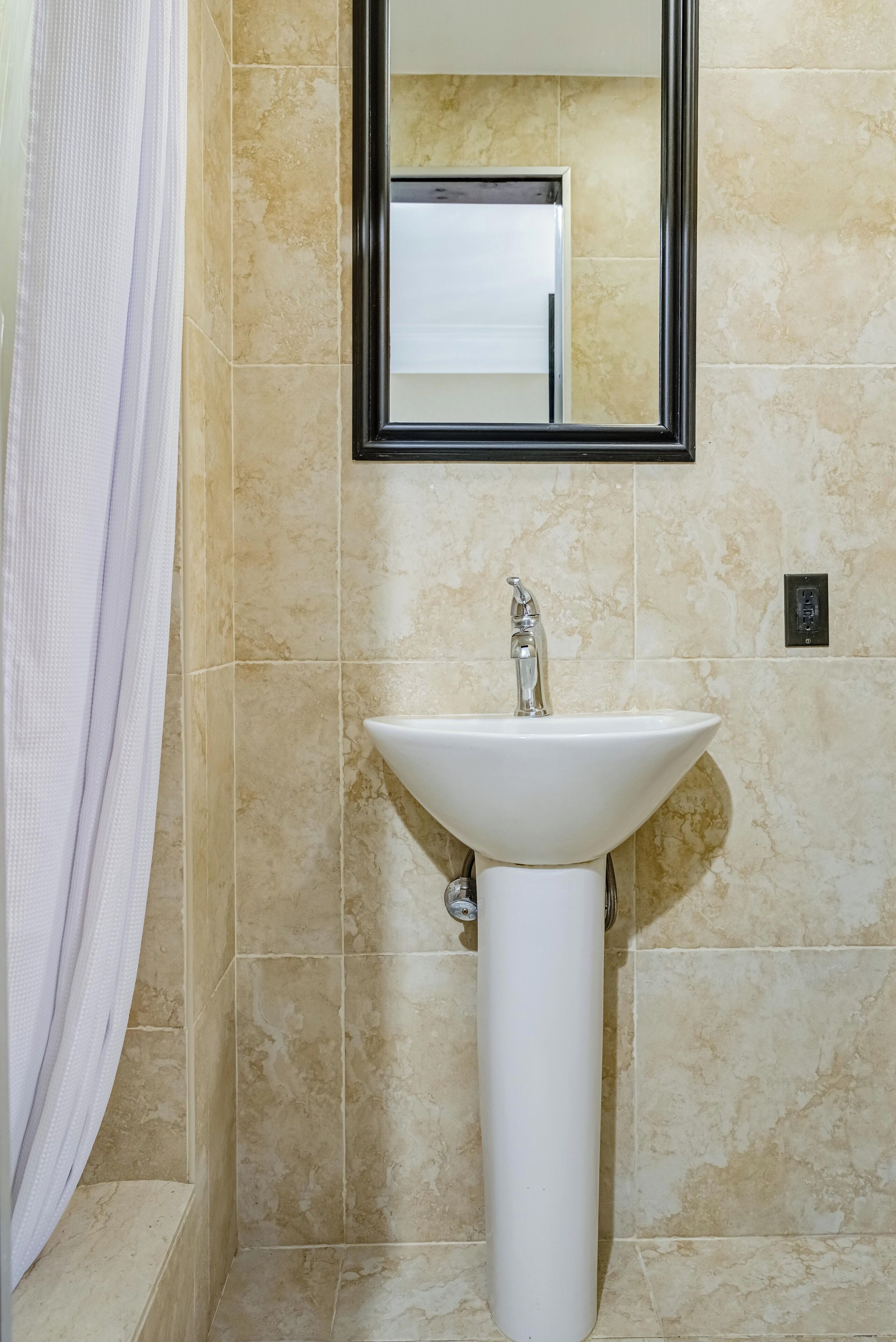 A bathroom with beige marble tiles, a white pedestal sink, a mirror with a black frame, and a towel rack.