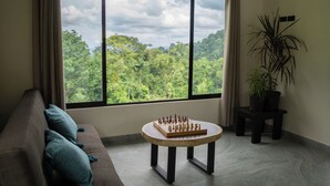 Una habitación con un ventanal grande, una mesa redonda de madera con un tablero de ajedrez, un sofá con cojines y una planta en maceta.