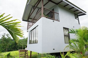 A two-story white building with a balcony, surrounded by lush greenery.