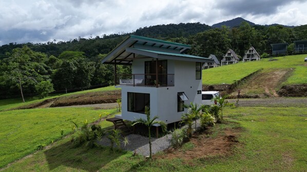 A two-story house with a balcony, surrounded by lush greenery and hills in the background.