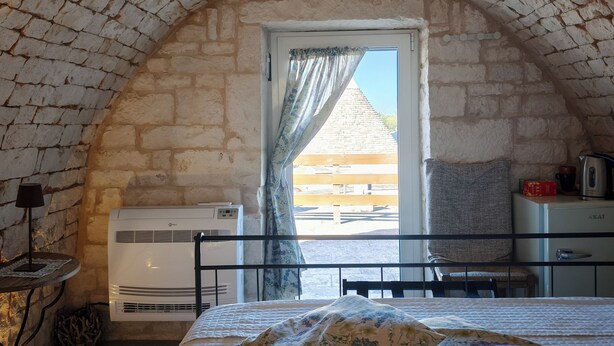 A bedroom with a stone vaulted ceiling, a bed with floral bedding, a window with curtains, and a view of a wooden deck outside. - High Accommodation in LocoMura (Locorotondo)
