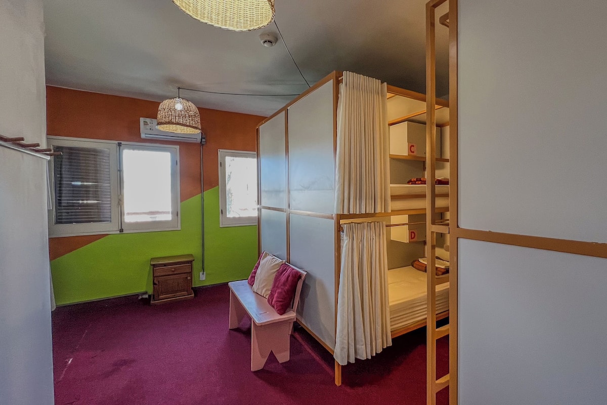 Een kamer met een stapelbed, een bureau en een stoel. Er zijn ramen en een plafondventilator.