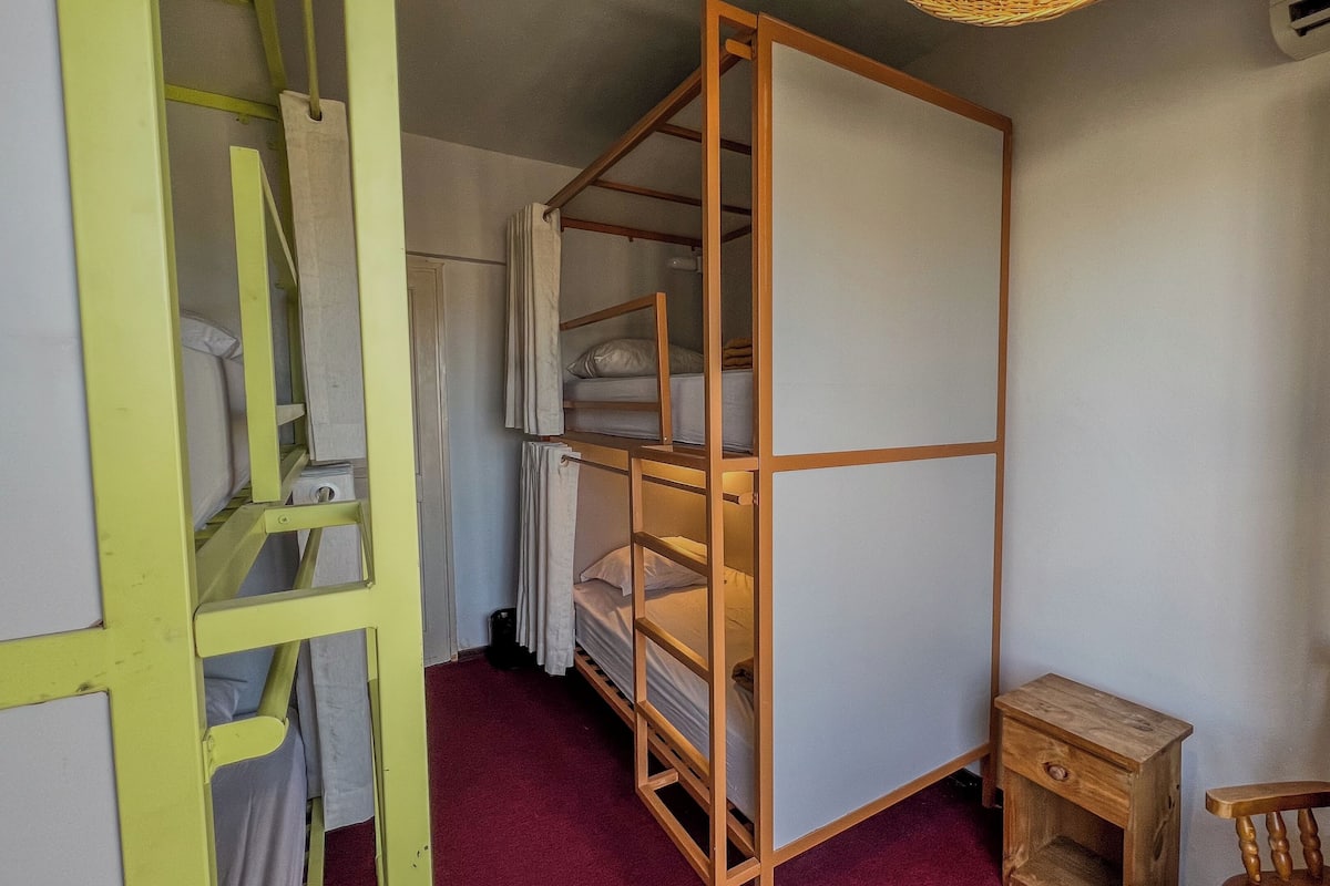 Een kleine kamer met een stapelbed, een klein houten nachtkastje en een stoel.