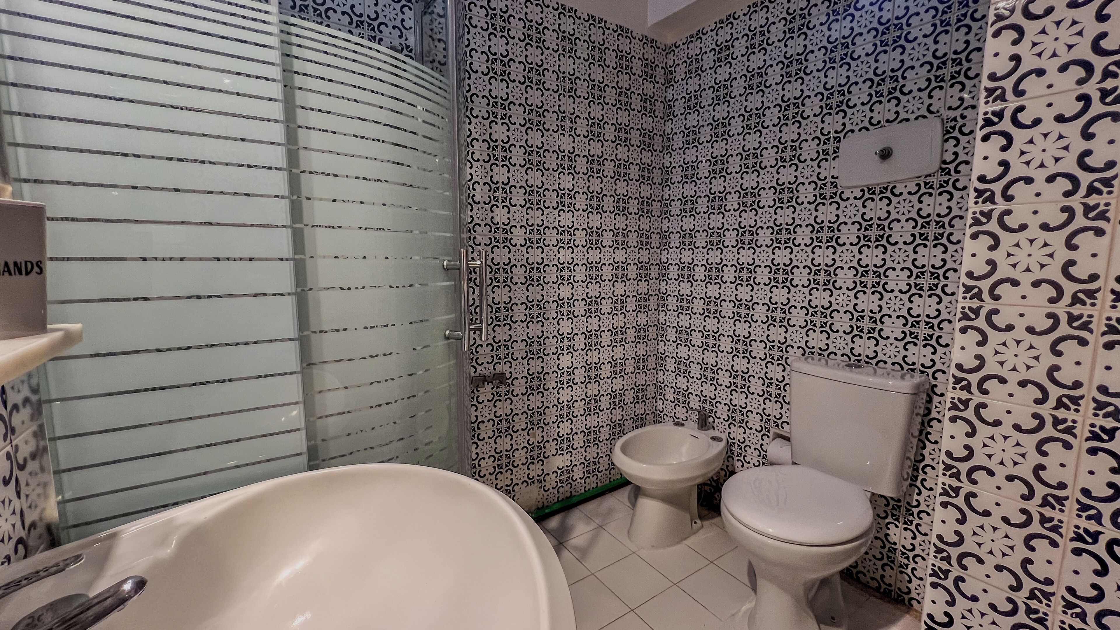Un baño con una bañera blanca, un inodoro blanco y un bidé. Las paredes están cubiertas con cerámica estampada en blanco y negro.