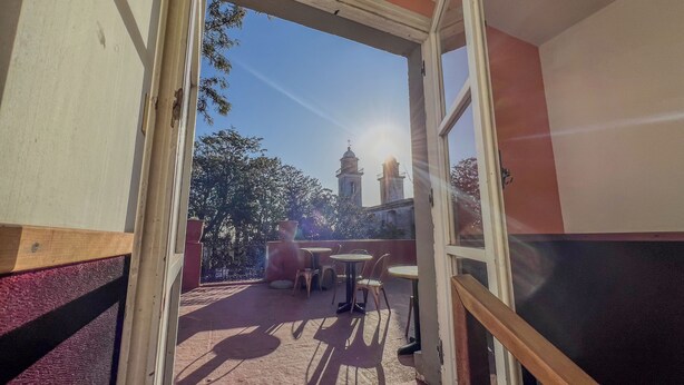 An open balcony with a view of a church tower, a table with chairs, and a potted plant.